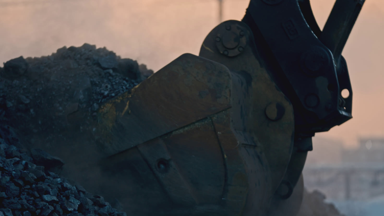 Close-up of an excavator bucket next to a heap of dark coal, in dusty evening light